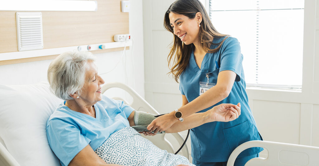 Nurse talking to senior woman in hospital bed. She is measuring her blood pressure.